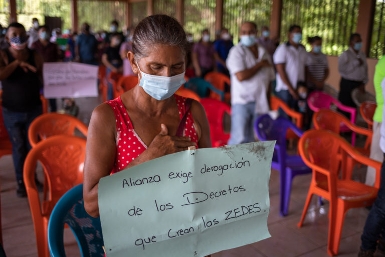 An Alianza resident sings the national anthem as she displays a sign opposing ZEDEs during the town hall meeting. Alianza, July 24, 2021. Photo by Martin Calix.